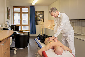 Orthopäde in weißem Kittel behandelt liegenden Patienten mit Tattoo auf dem Rücken in Praxisraum