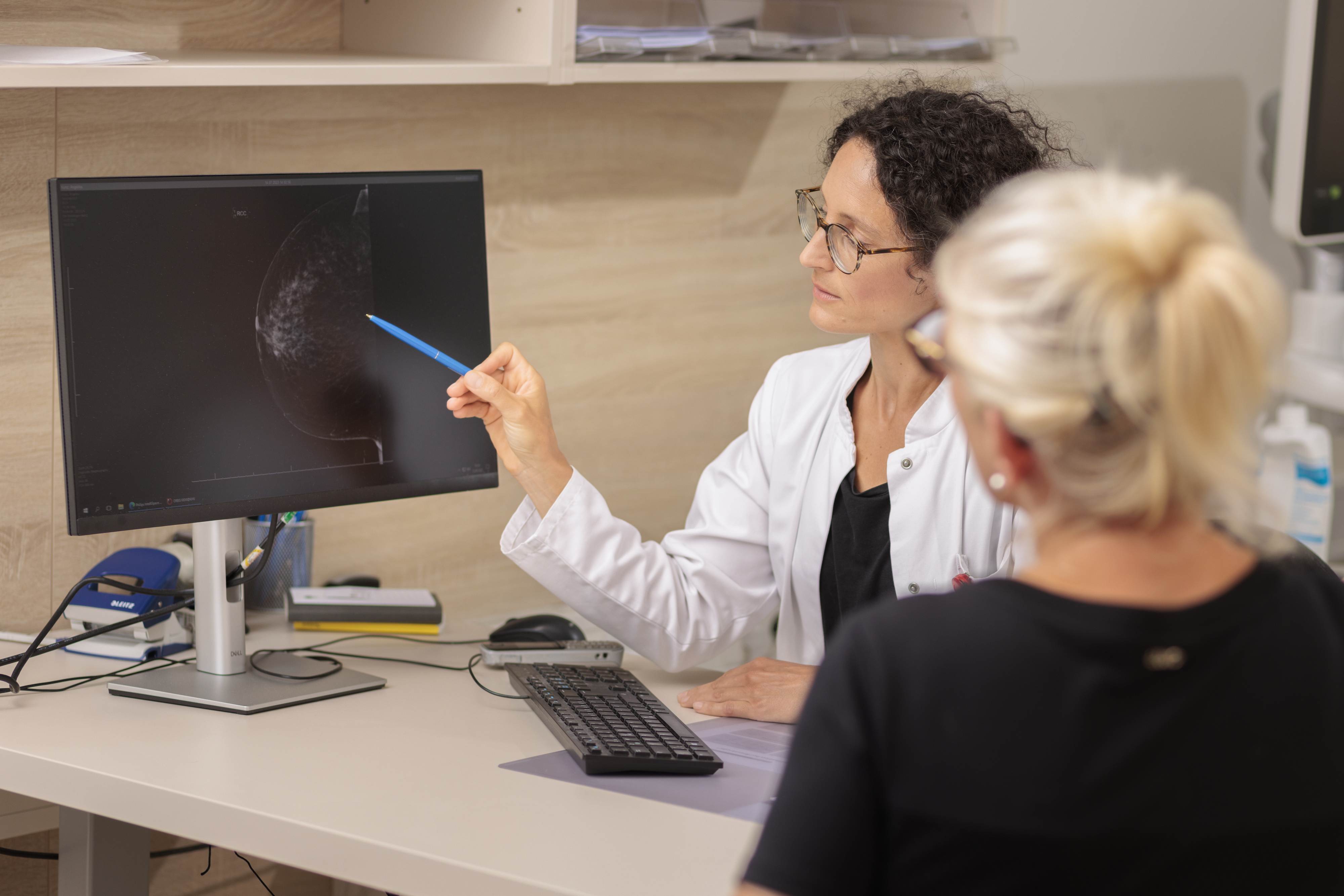Zwei Personen sitzen an einem Schreibtisch, eine zeigt mit einem Stift auf einen Computermonitor